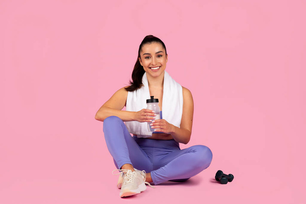 Una mujer en forma en ropa de ejercicio se sienta con una botella de agua y una toalla, con equipo de ejercicio sobre un fondo rosa - Foto, imagen
