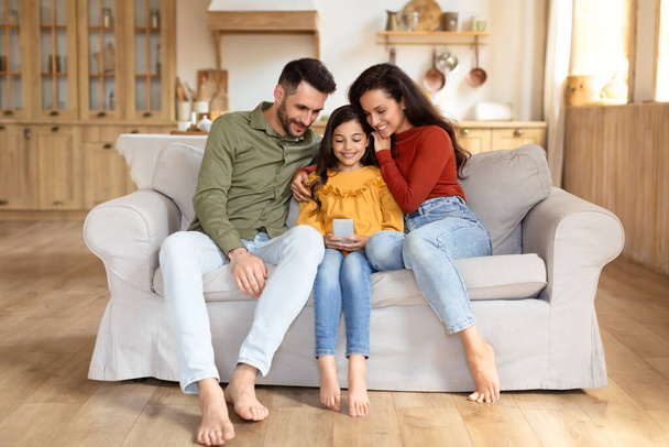 Μια ευτυχισμένη οικογένεια με μια κόρη που κοιτούν μια τηλεφωνική οθόνη μαζί, κάθονται σε ένα άνετο καναπέ σε ένα άνετο σαλόνι - Φωτογραφία, εικόνα