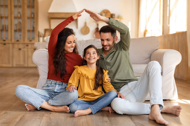 Hymyilevä perhe luo sydäntä lämmittävän kohtauksen muotoilemalla talon muodon käsillään istuessaan lattialla. - Valokuva, kuva