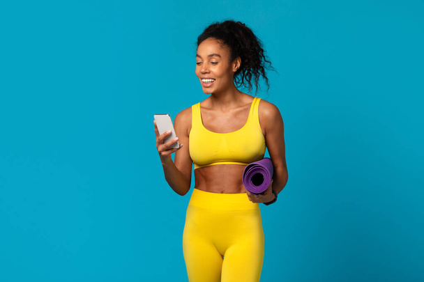 Cheerful African American fitness woman in yellow gym clothes holding a yoga mat and smartphone on a blue background - Photo, Image