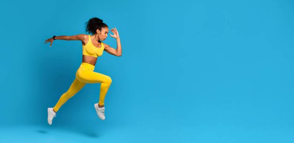Energetic young African American woman wearing yellow sports clothing captured in a dynamic mid-air jump on a blue background - Photo, Image