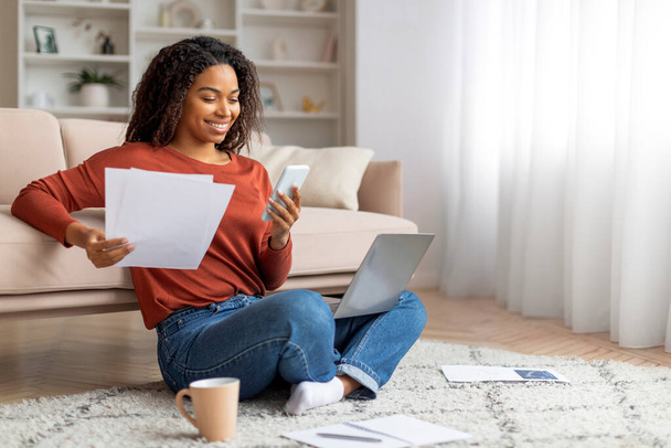 Весела чорна жінка, яка оглядає документи і тримає смартфон, сидячи на підлозі поруч зі своїм ноутбуком і чашкою кави, афроамериканка-фрілансерка вдома - Фото, зображення