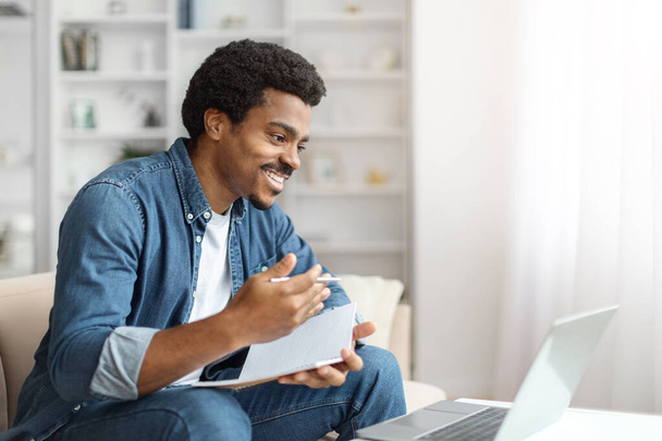 Een vrolijke zwarte man neemt deel aan een videoconferentie op zijn laptop, met notities, zittend op de bank in een goed verlichte kamer - Foto, afbeelding