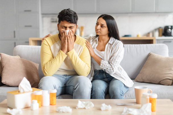 Indischer Mann pustet in die Nase, während eine Frau ihn auf dem Sofa tröstet, umgeben von Taschentüchern und Medikamenten. - Foto, Bild