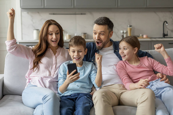 Radosna rodzina triumfalnie podnosi pięści patrząc na smartfona na kanapie w domu, rozrywka domowa - Zdjęcie, obraz