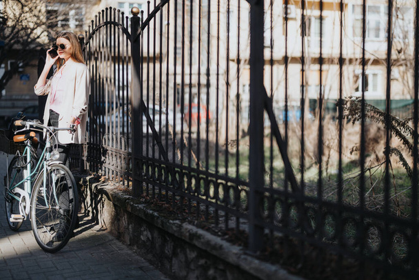 Mujer joven de moda en ropa casual usando su teléfono inteligente y apoyada en su bicicleta vintage por una valla de hierro forjado en un entorno urbano. - Foto, Imagen