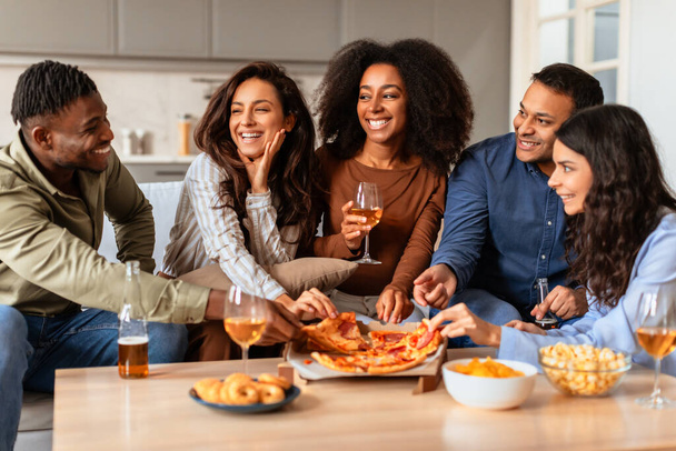 Різноманітна група молодих дорослих багаторасових друзів сміються разом з напоями та їжею під час комфортної домашньої вечірки - Фото, зображення