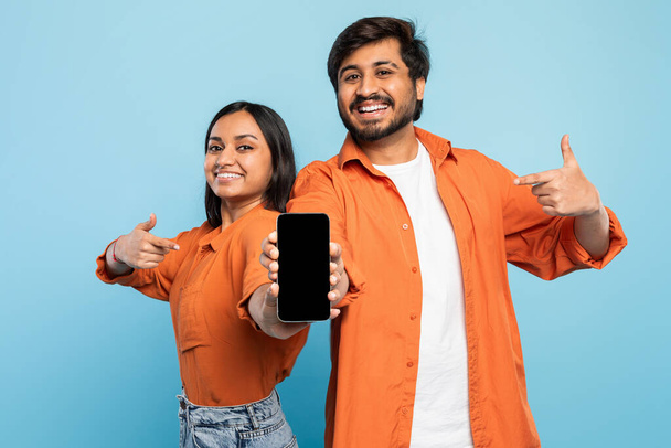 Para Indian uśmiecha się pewnie wskazując kciukiem na smartfona, idealny do reklamy technologii - Zdjęcie, obraz