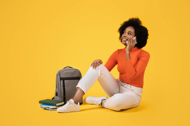 Thoughtful young woman sitting cross-legged, talking on the phone, dressed in an orange turtleneck, white pants, with books and a grey backpack beside her on a yellow background - Photo, Image