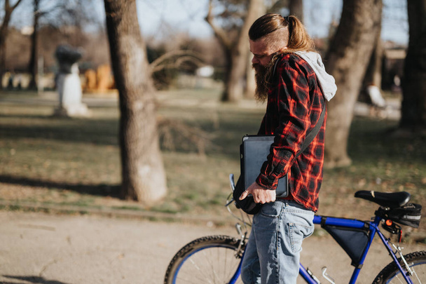 Ein bärtiger Geschäftsmann in Freizeitkleidung hält einen Laptop in der Hand, während er neben seinem Fahrrad in einem Stadtpark steht, ein Beispiel für Flexibilität bei der Arbeit aus der Ferne. - Foto, Bild