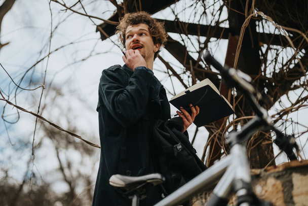 Contemplativo joven asociado de negocios de pie con la bicicleta en un entorno al aire libre, centrado en trabajar de forma remota con un portátil. - Foto, imagen