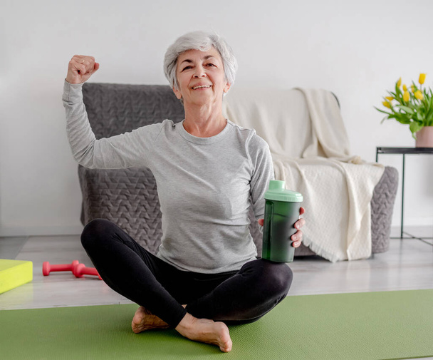Cheerful Elderly Woman Looks Into The Camera And Shows Off Her Bicep, Embodying An Active Lifestyle And Home Workouts - Foto, Imagem