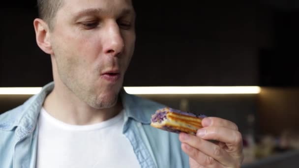 Young Caucasian Man eats an eclair in kitchen. A hungry man enjoys a delicious dessert. eating sweet donut unhealthy nutrition diet failure overeating obesity food addiction. - Footage, Video