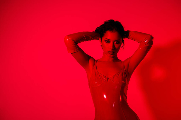 Фото ошеломляющей юной леди касаются волос сексуальная поза носить красную кожаную одежду изолированы на красном неоновом светлом фоне. - Фото, изображение