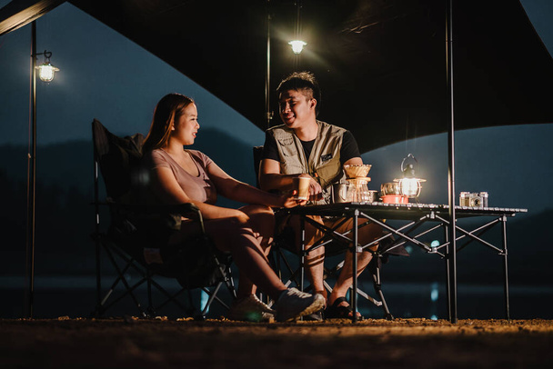 Pareja joven se sienta en una mesa de camping, charlando y relajándose debajo de una tienda de campaña al atardecer con una cálida luz de linterna. - Foto, Imagen