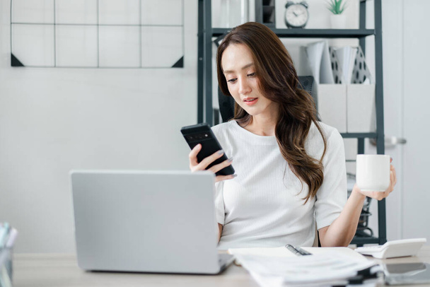 Geschäftsfrau checkt ihr Smartphone, während sie eine Tasse Kaffee in der Hand hält, einen Laptop offen auf dem Schreibtisch vor sich. - Foto, Bild