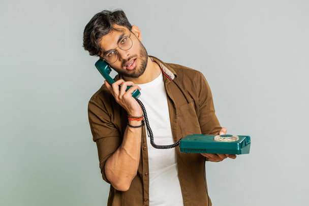 Tired bored Indian man talking on wired vintage telephone of 80s, fooling, making silly faces, exhausted of tedious story, not interested in communication talk. Arabian guy isolated on gray background - Photo, Image