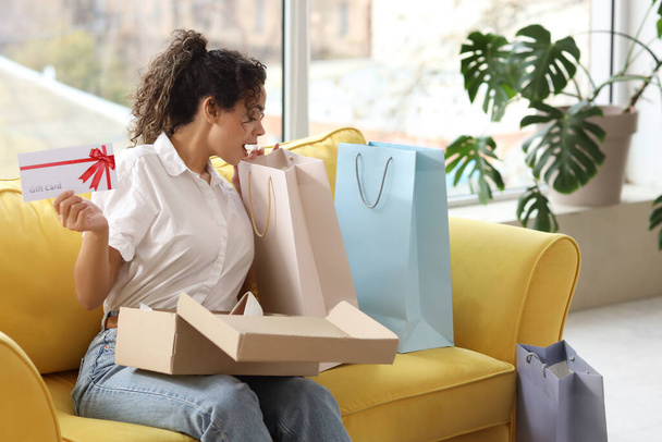 Młoda Afroamerykanka z kartą podarunkową i torbami na zakupy siedząca na kanapie w domu - Zdjęcie, obraz