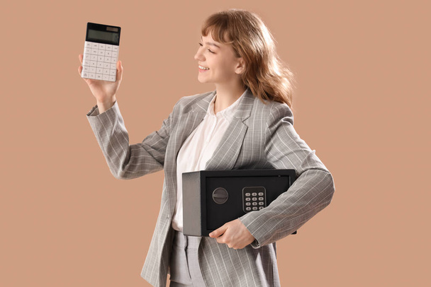 Mooie jonge gelukkige vrouw met kluis en rekenmachine op bruine achtergrond - Foto, afbeelding