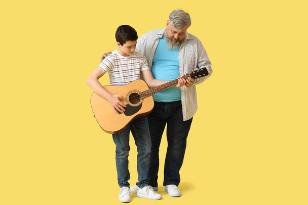 Χαρούμενος παππούς με τον χαριτωμένο εγγονό του να παίζει κιθάρα σε κίτρινο φόντο - Φωτογραφία, εικόνα