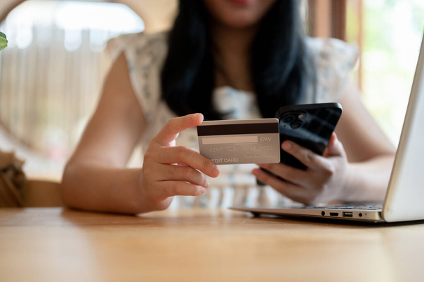A cropped image of an Asian woman holding a credit card and a smartphone while sitting at a table in a cafe. shopping online, transferring money, online payment, mobile banking - Photo, Image
