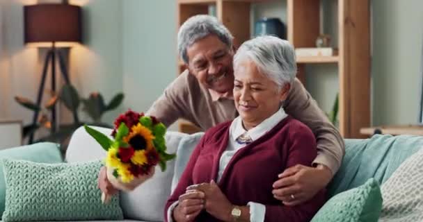 Couple, woman and old man with gift of flowers on sofa or couch, happy and living room of house. Senior, male person and partner with surprise for love, smile and happiness in home and retirement. - Footage, Video