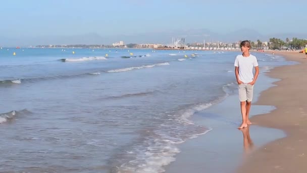 ホワイトTシャツのハンサムなティーンエイジャー少年は,夏の週末の休暇を楽しむ海岸線に沿って歩きます, サルー市スペイン. 高品質のフルHD映像 - 映像、動画