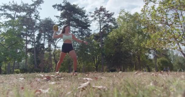 Une femme sportive jouit d'un match de balle lors d'une journée ensoleillée dans un parc rempli de grands arbres et de feuilles éparses. - Séquence, vidéo