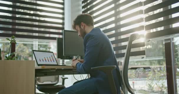 Een toegewijde mannelijke zakenman richt zich op business analytics, het beoordelen van kleurrijke grafieken op een laptop in een goed verlichte kantooromgeving, een voorbeeld van ijver en professionaliteit. - Video