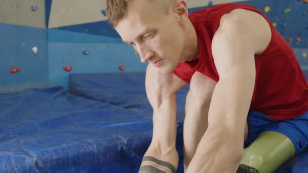 Tilt down shot of male athlete putting shoe on prothetic leg before training in climbing gym - Footage, Video