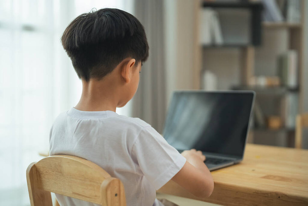 Młody chłopak siedzi przy stole z laptopem przed sobą. Skupia się na pracy, prawdopodobnie odrabianiu lekcji lub nauce. Koncepcja koncentracji i determinacji, gdy chłopiec pracuje nad swoim zadaniem - Zdjęcie, obraz