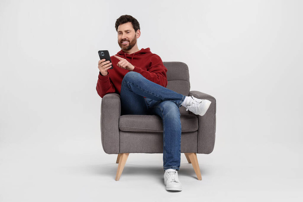 Happy man with smartphone sitting on armchair against white background - 写真・画像