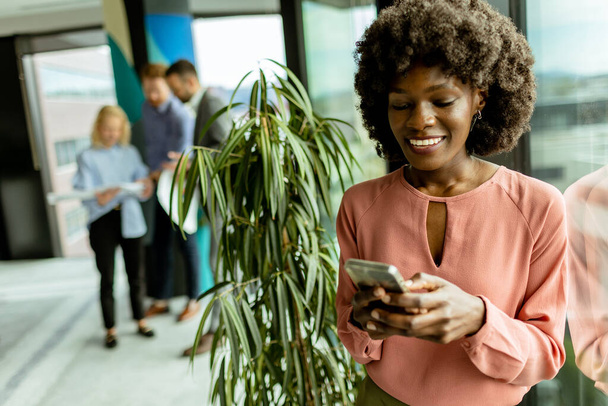 Afrikkalainen amerikkalainen nainen hymyilee puhuessaan puhelimessa työtovereiden keskellä toimistoympäristössä, joka huokuu ammattimaisuutta ja iloa.. - Valokuva, kuva