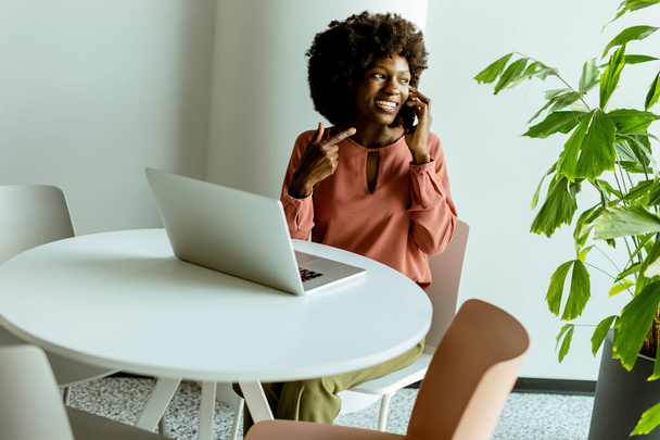 Een vrolijke vrouw praat op haar telefoon aan een cafe tafel met een laptop en een levendige plant naast haar. - Foto, afbeelding