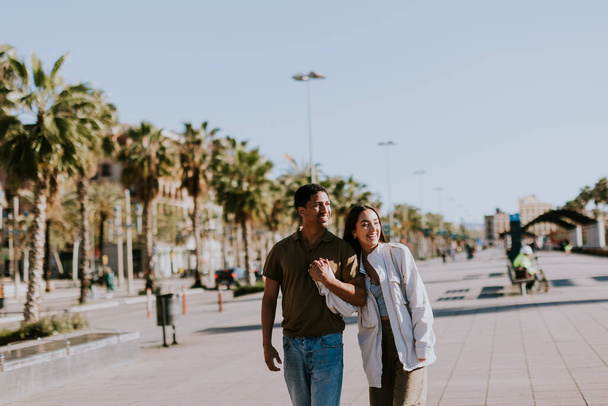 Młoda para trzyma się za ręce i uśmiecha idąc słoneczną ścieżką w Barcelonie, oprawioną dłońmi - Zdjęcie, obraz