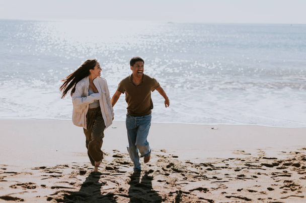 笑顔のカップルは,砂浜の海岸で気取らない走りを楽しみ,バックグラウンドで穏やかに壊れる波 - 写真・画像