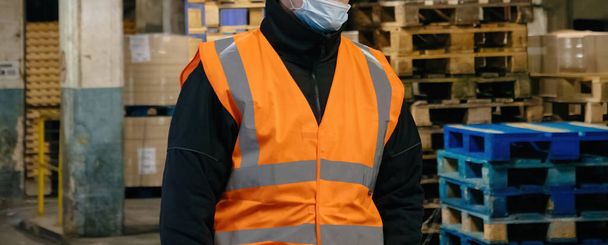 Nerozpoznatelný pracovník v dobře viditelné vestě a masce stojí bdělý ve skladišti. - Fotografie, Obrázek