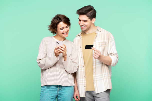 Muotokuva hymyilevä pari, houkutteleva mies ja nainen seisoo eristetty turkoosi tausta, tilalla matkapuhelin, mobiilisovelluksella, tilalla luottokortti, maksu. Verkkokaupan käsite  - Valokuva, kuva