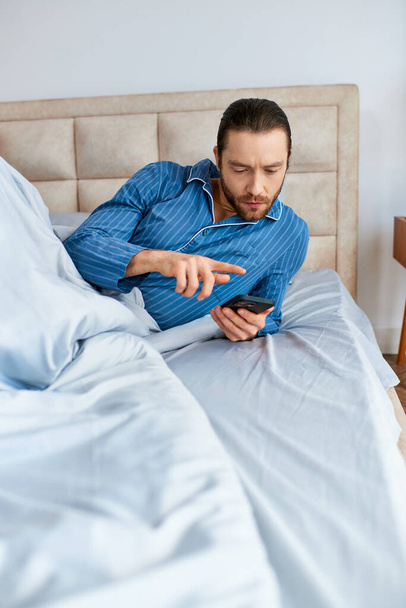 Een man ligt in bed, geabsorbeerd door zijn mobiele telefoon scherm. - Foto, afbeelding