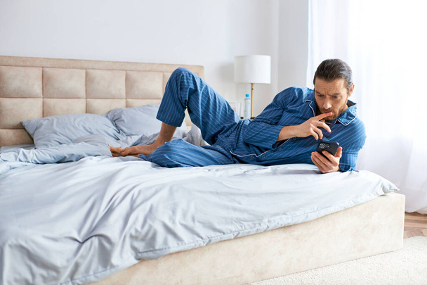 Красивый мужчина лежит на кровати, практикует йогу, держа в руках мобильный телефон.. - Фото, изображение