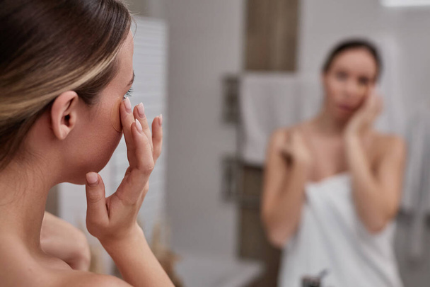 Κλείσιμο της νεαρής γυναίκας χρησιμοποιώντας κρέμα ματιών στο μπάνιο και κοιτάζοντας στον καθρέφτη απολαμβάνοντας την ημέρα φροντίδας στο σπίτι, αντίγραφο χώρο - Φωτογραφία, εικόνα