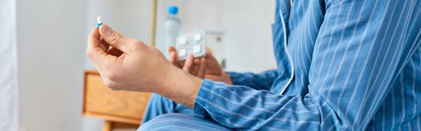Un hombre se sienta en una cama, sosteniendo una botella de agua y una pastilla. - Foto, imagen