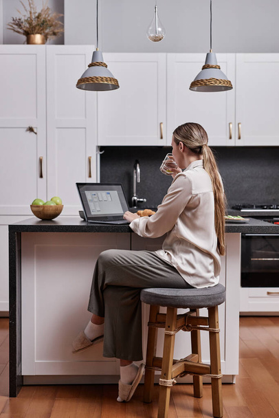 Vertical full length portrait of young woman using laptop at kitchen table in home interior - Photo, Image