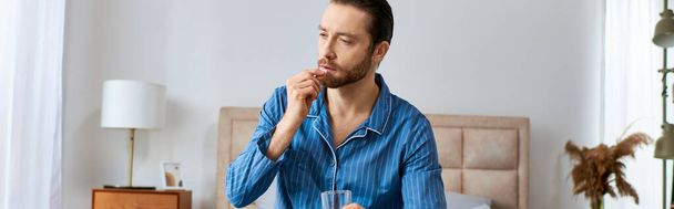 Ένας άντρας κάθεται σε ένα τραπέζι με ένα ποτήρι νερό μπροστά του και παίρνει χάπια.. - Φωτογραφία, εικόνα