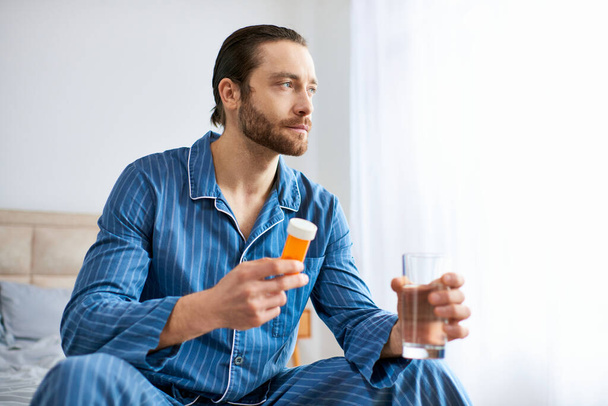Όμορφος άντρας κάθεται στο κρεβάτι, κρατώντας ένα μπουκάλι χάπια και γυαλί. - Φωτογραφία, εικόνα
