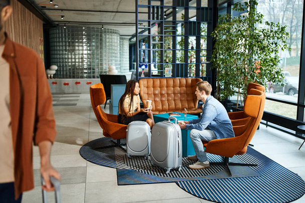 Homem e mulher multicultural sentados em um sofá, vestindo roupas casuais, em um lobby do hotel durante uma viagem corporativa. - Foto, Imagem