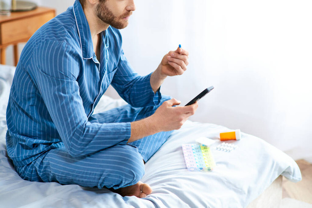 Um homem relaxa em uma cama, segurando pílulas e um celular. - Foto, Imagem