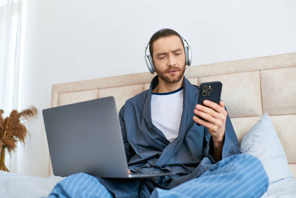 Ένας άνθρωπος απολαμβάνει το πρωί, χρησιμοποιώντας ένα φορητό υπολογιστή και ακουστικά σε ένα κρεβάτι. - Φωτογραφία, εικόνα