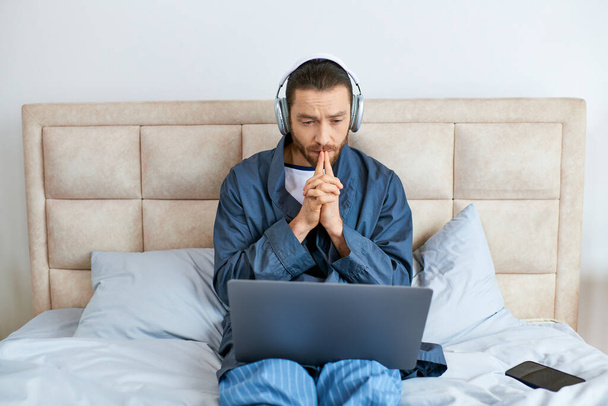 Un hombre sentado en una cama, usando auriculares, usando una computadora portátil. - Foto, imagen