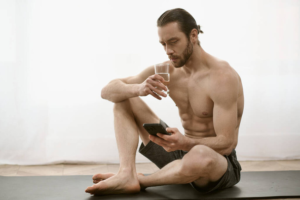 Shirless man zittend op yoga mat, starend naar mobiele telefoon. - Foto, afbeelding